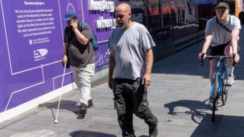 A man walks down the pavement in London with a white cane