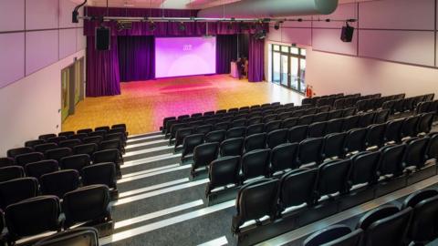 A new hall with tiered seating, a curtained off area for a stage, a large screen, lectern and apparatus on the ceiling for lighting and sound.