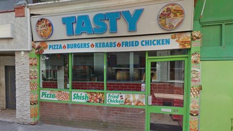 Street view of the Tasty takeaway. It has a blue sign on a white background that says "pizzas burgers kebabs fried chicken" underneath with glass windows advertising pizza, shish and fried chicken