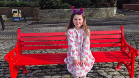 Ivy sits on a bright red bench on a cobbled street, she wears a white dress with pink love hearts on it and has a headband with purple tinsel and hearts on