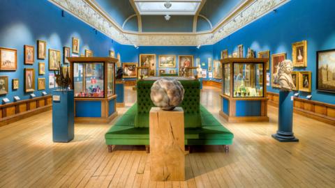 The new Upper Gallery space with artworks and sculptures and a green sofa in the middle. The walls have been painted a bright royal blue.