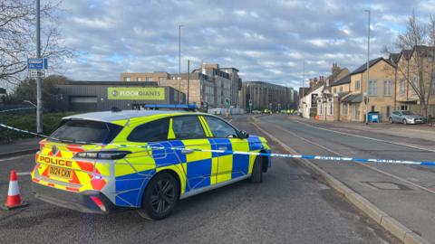 The rear of a police car is shown inside police tape on a road. In the distance, more police cars and officers can be seen