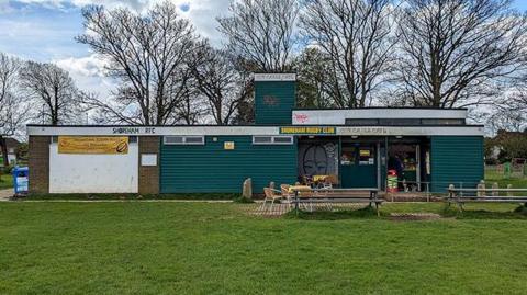 A single-storey green pavilion at the top of a green grass bank. In front of the building are picnic benches, behind the building are trees with no leaves