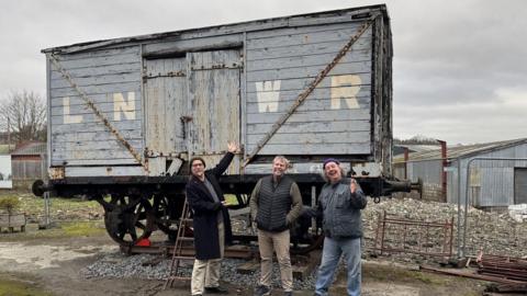 Three men are standing in front of an old railway wagon. It blue-grey and wooden, with the letters "LNWR" painted on the side in white. It is siting on a small piece of track on some stones.