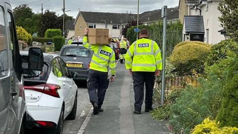 Workers in yellow hi vis jackets walking down a residential street in a cloudy day. They are facing away from the camera. One of them is carrying a cardboard box. More workers could be seen in the distance ahead. Some vehicles are parked on the side of the road.