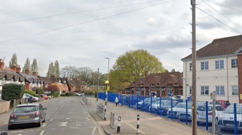 A Google Streetview image of West Byfleet infant and junior schools, a white and red building behind a blue fence, and a faded zebra crossing on the road in front. 