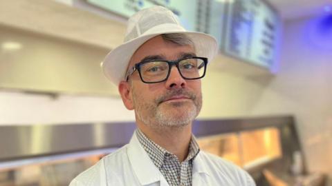 Marco Valente looking straight at the camera. He is wearing a white hat and has glasses. He is standing in front of the counter at his fish and chip shop.