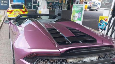A purple Lamborghini parked next to the fuel pumps at a petrol station. In front of the sports car is a parked marked fluorescent yellow and orange police car. The vehicle says POLICE on the back. 