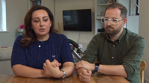 A man, Tom Hender, wearing a dark green shirt, white and red glasses and a watch, and a woman, Ewa Hender, wearing a dark blue top with a necklace and a watch, sit at a wooden table. There are shelves, a television and other furniture in the background.