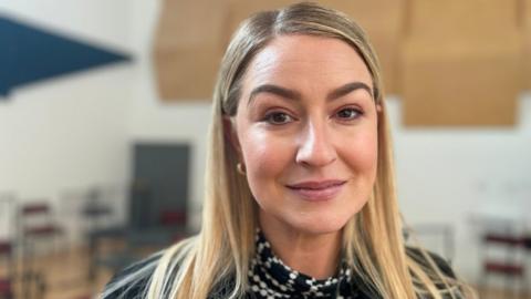 Imogen has long, blonde hair and is wearing a black and white patterned blouse with a black blazer. She is stood in a musical rehearsal space.