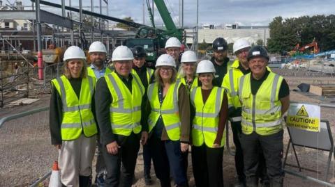 A group of of 11 people stand in florescent yellow jackets and hard hats. Behind them the construction site can be seen. There is a sign next to them that says "Caution deep excavations"