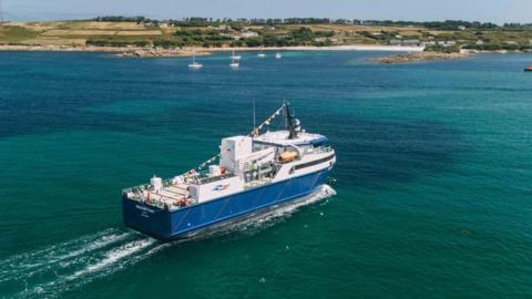 A large blue vessel heads towards the Isles of Scilly across blue waters with boats and land seen in the background 