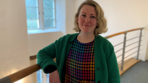 Woman in green jumper stood in a hallway with right arm on a railing