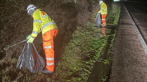 Two people in hi-vis clothing, helmets and gloves and holding large black rubbish bags and sticks, on a verge by the side of a road.