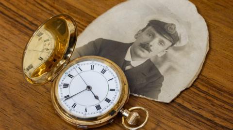 A gold pocket watch and an old black and white photo of a man with a moustache wearing nautical uniform. The items are both placed on a wooden surface.