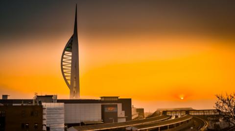 The sky glows orange over the city of Portsmouth. It is a glowing ball appearing through the cloud. The train station is in the foreground with a cinema complex and the white Spinnaker Tower behind