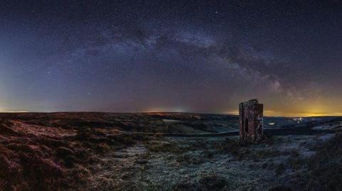 Dark skies above Rosedale, in the North York Moors. A landscape image, of moorland with the ruins of a tower to the left. Clusters of stars can be seen in the sky, which is a purple hue.