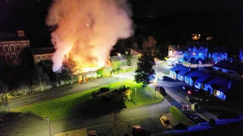 An aerial photo of Sinton's Mill in Tandragee on fire on Tuesday night.  A large plume of smoke and bright orange flames are rising from the Victorian brick complex which is in a housing estate.  Firefighters hoses are stretched across the green in front of the mill.  Houses and cars are in the foreground. 