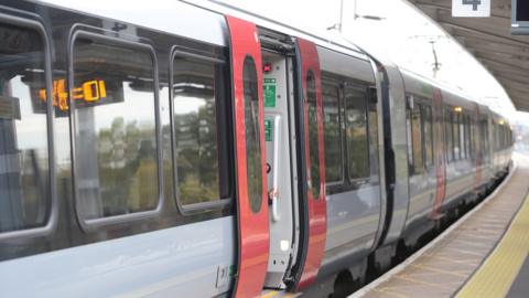 Image is a train stopped on a platform with one of the doors open. It is a Greater Anglia Service and follows a light grey and red colour scheme.