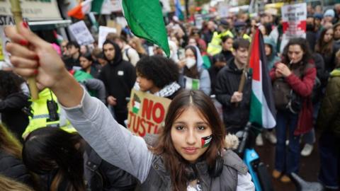A young woman looks at the camera while waving a flag, as crowds of protesters walk behind her
