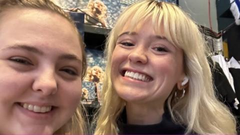 Hannah Clayton on the left having a selfie with Maisie Peters who is to to her right in the record store. It is a close-up photo of both women who are smiling for the camera. Hannah has brunette hair and Maisie has blonde hair and white airpods in her ears