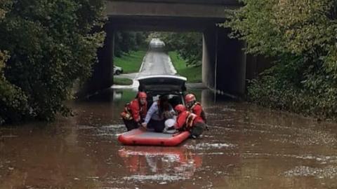 A woman is helped out the back of a car surrounded by water by firefighters