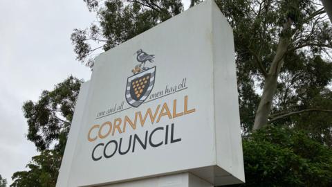 A large white sign with the words 'Cornwall Council'. There is a badge above the text. Behind the sign are trees.