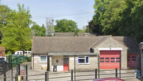 The exterior of Whaley Bridge fire station - a single storey building with two red shutters, a red front door and two white framed windows. The building is surrounded by a black metal fence.