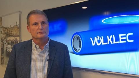 Phil Popham, executive director at Volklec, stands in front of a television displaying the company's logo. He is wearing a blue suit jacket and a white pinstripe shirt with an open collar.