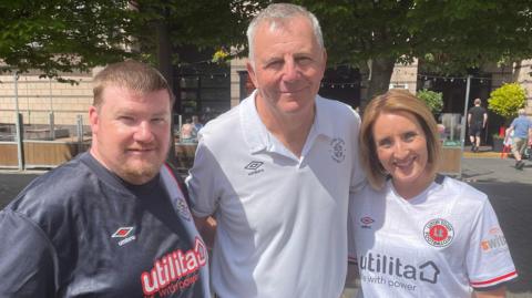 Left to right: Kevin, Paul and Helen stand together in a group, smiling at the camera, in St George's Square in Luton. 