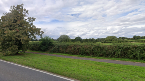 A field, with a tree to the left and hedgerow in front off it and then many trees in the background into open countryside, to the east of Ploughley Road in Ambrosden, taken from the road by Google 