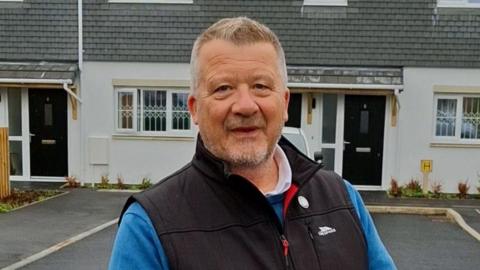A man smiles at the camera while stood in front of a residential house wearing a black gilet over a blue jumper