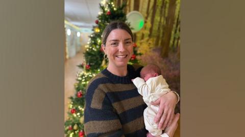 Chloe and her newborn son Alfie pictured in front of a Christmas tree, a short time after his birth on Christmas Day 2024. Chloe is wearing a blue and brown striped jumper and is holding Alfie on her left side. Alfie is wearing a cream Babygro. 