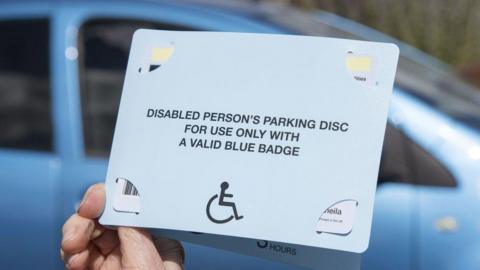 A hand holding a blue badge. There is a blue car in the background. 