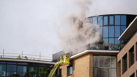 A plume of smoke comes out of a building, with firefighters on scene. 