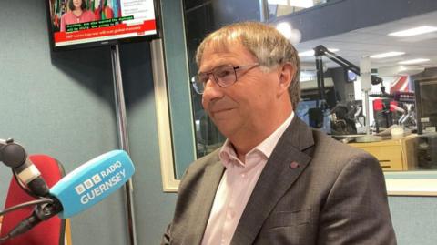 A man wearing a grey suit, pink shirt and glasses. He is looking to the left. In front of him is a BBC Radio Guernsey Microphone. Behind him is a blue wall and large window.