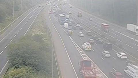 A traffic cam showing a motorway at a junction. The road is lined with thick woodland. On the left, a slip road joins the main motorway which is heavy with traffic moving away from the camera. The other side of the central reservation contains lighter traffic and a white lorry is leaving via a slip road which disappears from view.