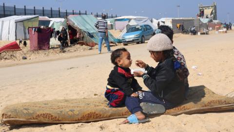 Displaced Palestinian girl feeds her brother at a tent camp in Rafah