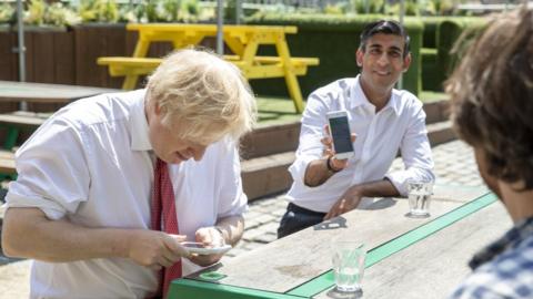 Prime Minister Boris Johnson (L) and Britain's Chancellor of the Exchequer Rishi Sunak (R) use their smartphones as they visit Pizza Pilgrims in West India Quay, London Docklands on June 26, 2020