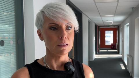 Emma Wilding gives a TV interview in a corridor. She has short, dyed white hair in a stylish short side sweep and she wears make-up and a black dress.
