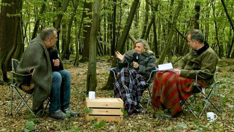 Three people sit in the woods on camping chairs