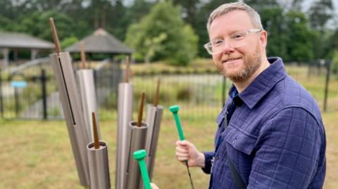 Stuart Wood stands next to a set of tubular bells with a pond behind him while holding two chime hammers.