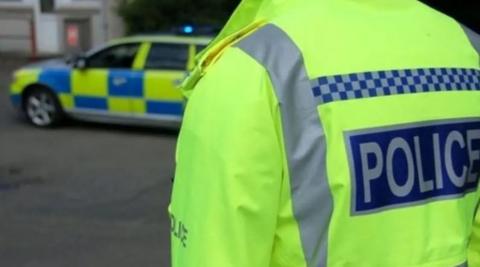 Back of a police officer, wearing a fluorescent jacket that reads Police. There is also a police car in the background. 