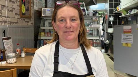 Helen Lawson has shoulder length brown hair and a white T-shirt with a black apron over it.
