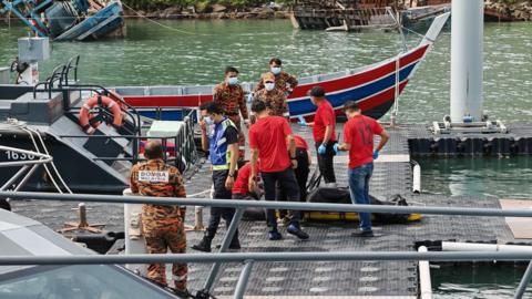 Men in red shirts and green/orange camouflage attire standing around a black body bag lying on a stretcher.