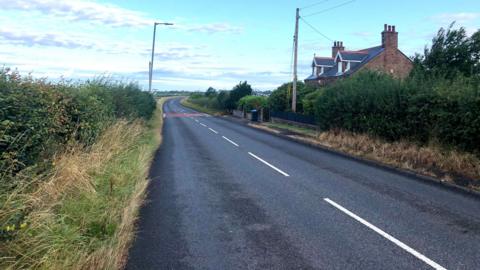 A long straight on a rural road with a bend to the right at the end. There are hedgerows up either side and a house to the right.