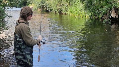 An angler holding a fishing rod and wearing waders stands in the River Tone