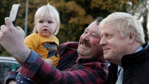 A father taking a selfie of his son and Boris Johnson during the PM's visit to a railway station near Manchester