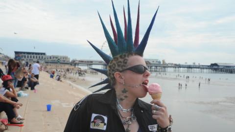 A punk with multicoloured spiked hair licks an ice cream on the Blackpool promenade. A pier is in the background.