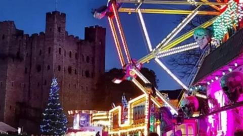 A lit Christmas tree and colourful lit fairground with big wheel in foreground with Rochester Castle in the background. 